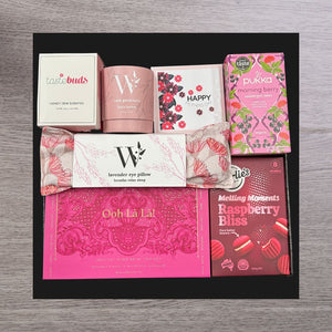 Gift box with pink-themed items: a honey dew candle, rose geranium bath bomb, Mother's Day card, tea box, lavender eye pillow, hand balm set, and raspberry cookies.