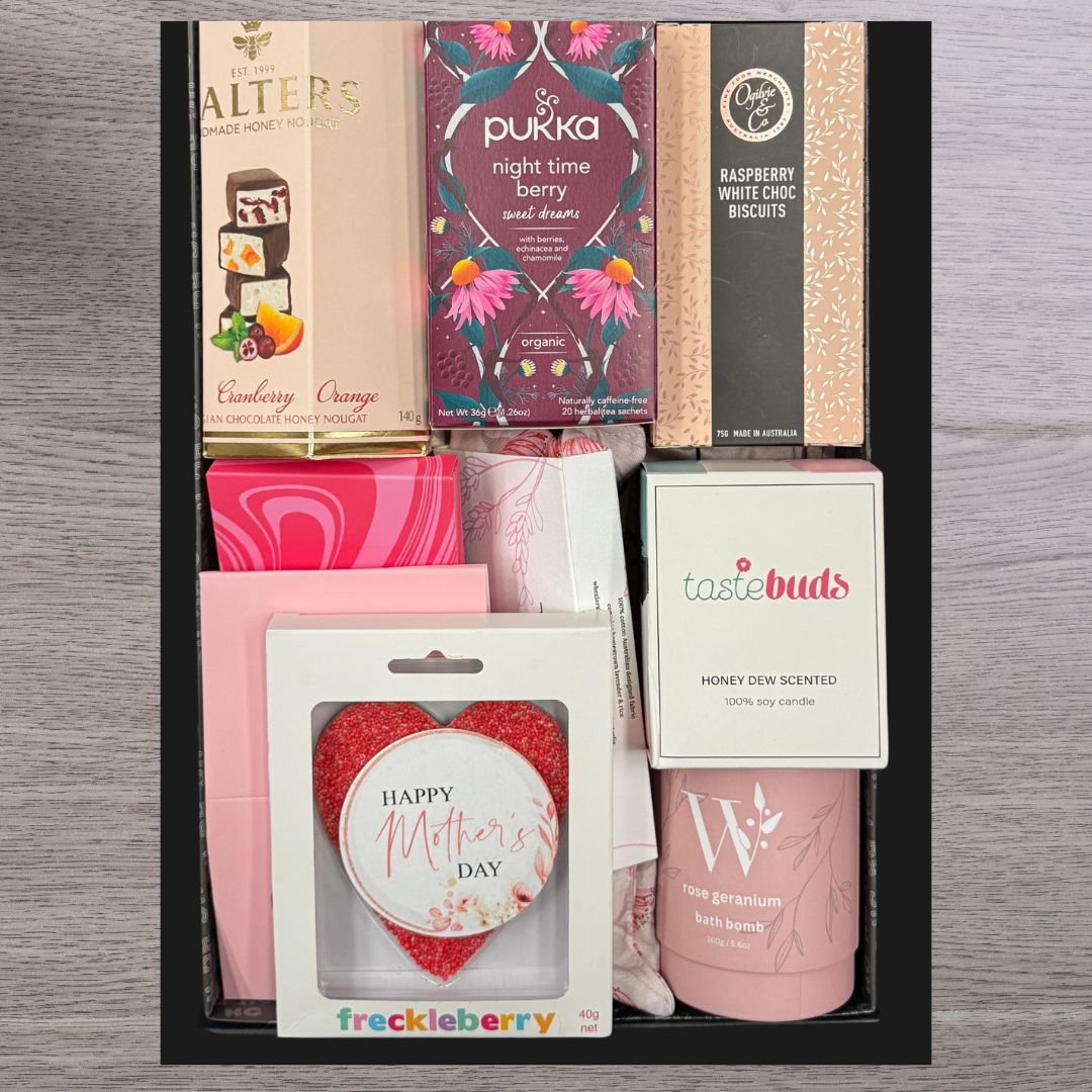 Gift box with various items including tea, chocolate, biscuits, and heart-shaped "Happy Mother's Day" treat. Soft pink tones, elegant packaging.