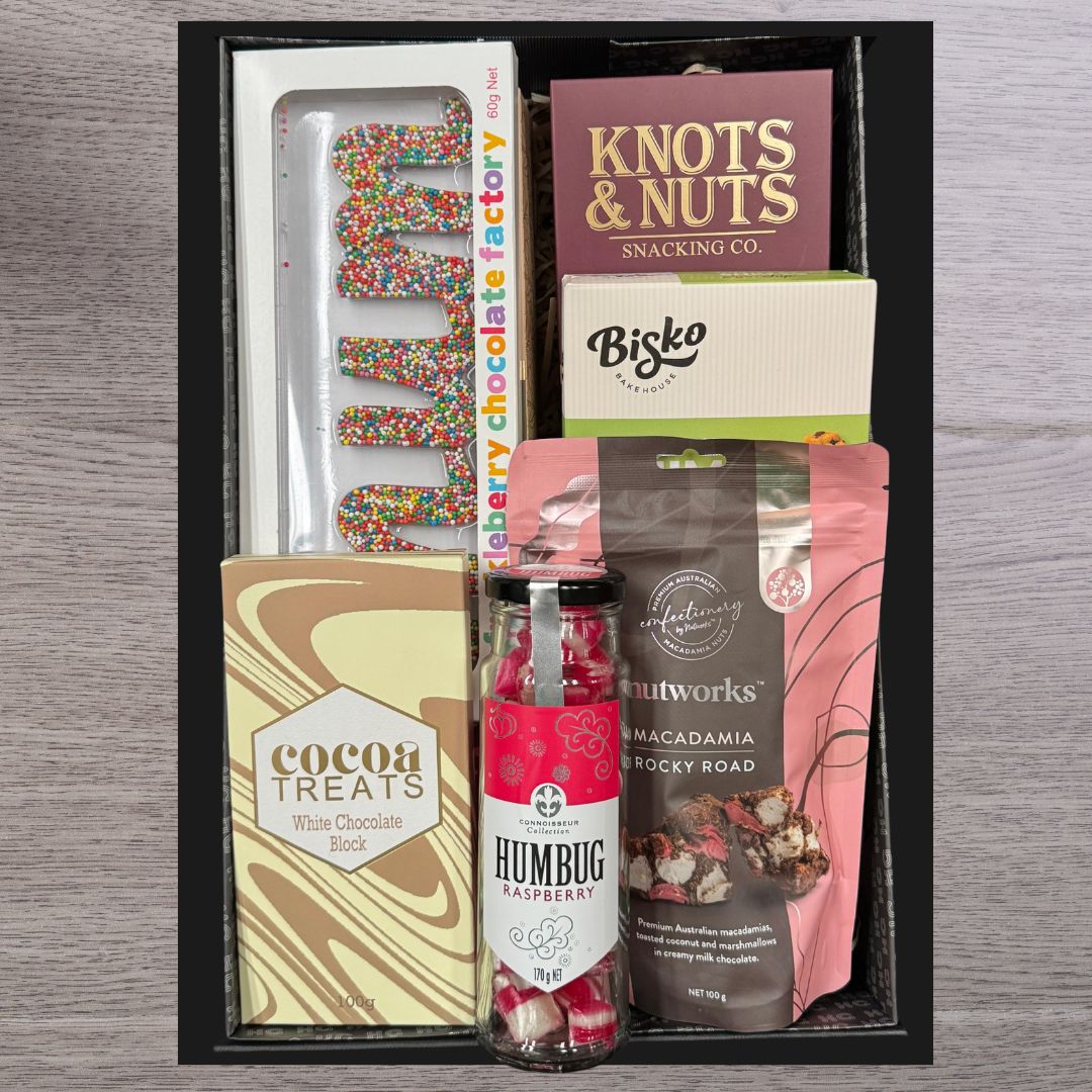 Gift box on a wooden table containing colorful "YUM" letters, Knots & Nuts pack, cocoa treats, Bisko pack, raspberry humbugs, and macadamia rocky road.