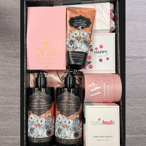 A gift box on a wooden surface contains various pampering items, including pink bath salts, floral body cream, hand and body wash bottles, and a “Happy Mother’s Day” card, evoking a serene and celebratory tone.