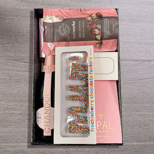 Gift box with a pink Chandon bottle, macadamia rocky road, chocolate letters covered in colorful sprinkles, and bath salts, on a wooden table.