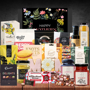 Mother's Day gift collection with various gourmet treats. Includes snacks, chocolates, jams, and a floral card, arranged on a wooden surface.