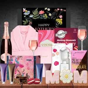 A festive Mother's Day gift display featuring a pink robe, rosé champagne, floral-themed chocolates, beauty products, a journal, and decorative cookies spelling "MOM."